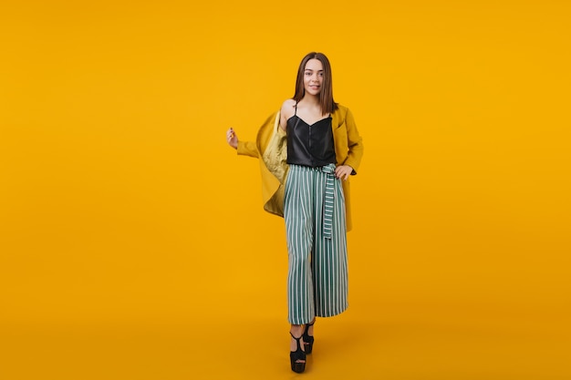 Full-length portrait of slim stylish girl with brown hair standing. Indoor photo of blissful white lady in elegant striped pants.