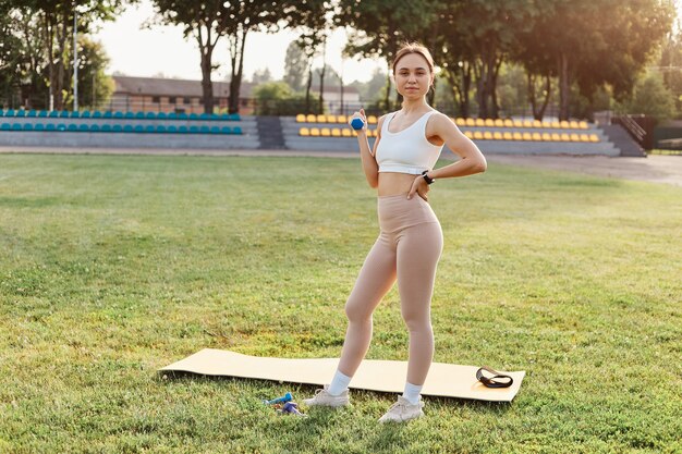 Full length portrait of slim woman with perfect body, brunette girl wearing beige leggins and white top looking at camera and holding dumbbell, training biceps and triceps in stadium.