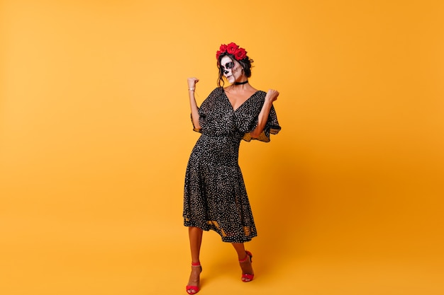 Full-length portrait of slim woman with roses in hair celebrating day of the dead. Gorgeous girl in mexican party outfit dancing on yellow background.