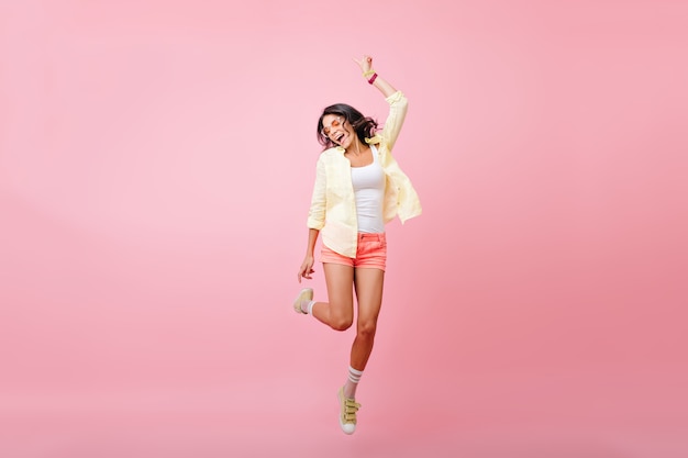 Full-length portrait of slim young woman with tanned skin jumping with smile. portrait of happy brunette girl in yellow shirt, dancing and singing with eyes closed.