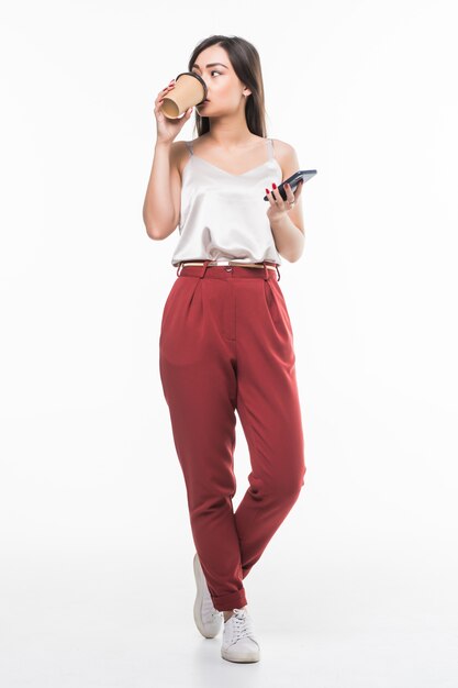 Full length portrait of a smiling asian businesswoman using mobile phone while holding cup of coffee to go isolated over white wall