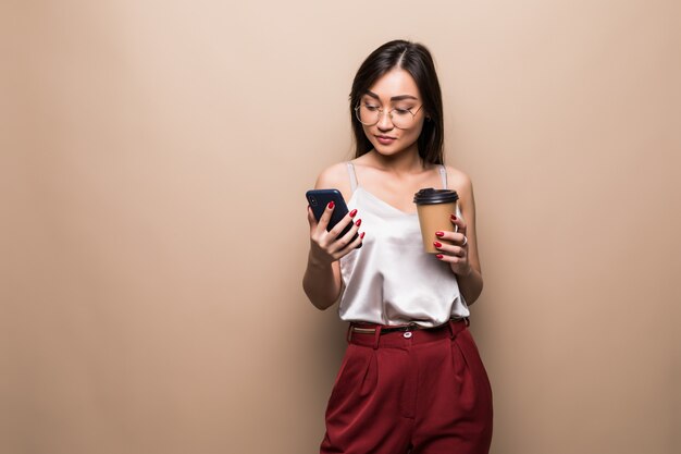 베이지 색 벽 위에 격리 이동 커피 잔을 들고 휴대 전화를 사용하여 웃는 아시아 여자의 전체 길이 초상화
