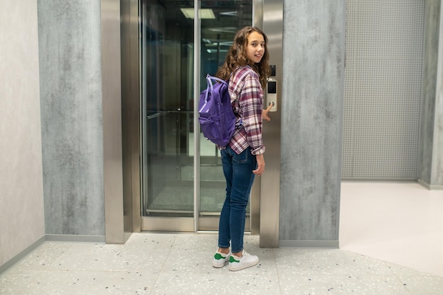 Full-length portrait of a smiling calm kid standing before the elevator door and pressing the call button