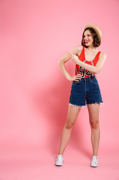 Full length portrait of smiling teen girl in red bodysuit and jeans short standing with hand on her waist, pointing with finger, looking aside