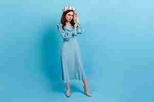 Free photo full-length portrait of sophisticated lady in midi dress. woman with crown of flowers on blue wall.