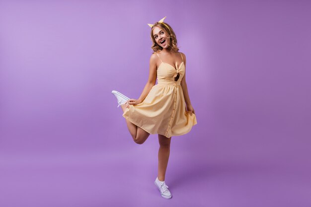 片足で立っている見事な日焼けした女の子の全身像。紫で踊る黄色のかわいいドレスを着たポジティブなスリムな女性。