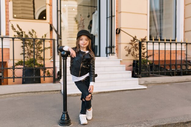 Full-length portrait of stylish kid standing with legs crossed next to iron pillar in front of shop.