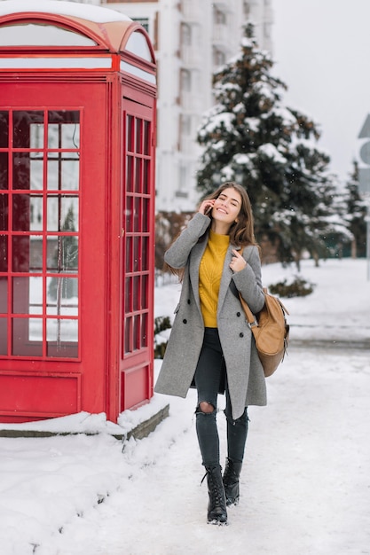 Ritratto a figura intera di giovane donna alla moda in cappotto grigio e pantaloni strappati parlando al telefono, camminando per la strada innevata. foto della splendida donna in piedi vicino alla cabina telefonica rossa e tenendo lo smartphone.