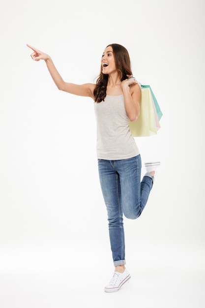 Full-length portrait of trendy woman pointing finger away and holding many shopping bags, isolated over white wall