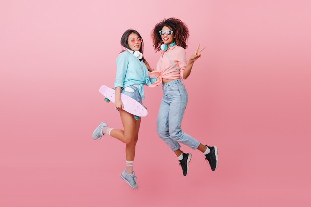 Full-length portrait of two sporty ladies jumping and smiling. Glamorous skater girl in blue shirt having fun with african female friend in black shoes.