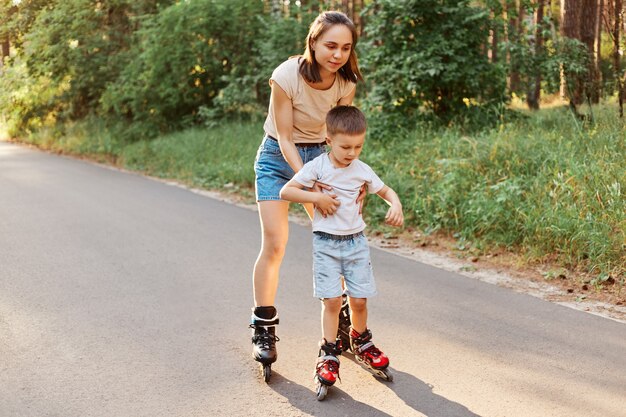 女性と幼い息子が一緒にローラーブレードをしている様子、母親が子供にローラースケートを教えている様子、かわいい男の子がローラースケートに乗って学んでいる様子の全身像。