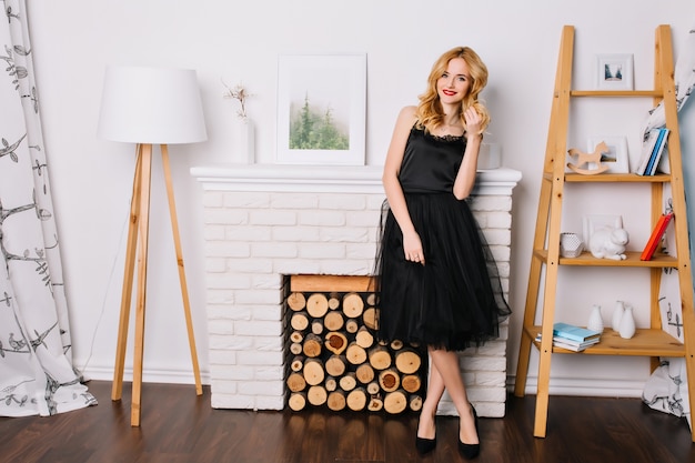 Full length portrait of young blonde woman in bright room with nice, modern interior, floor lamp, fake fireplace, shelving with figurines, books. Wearing stylish black dress and shoes.