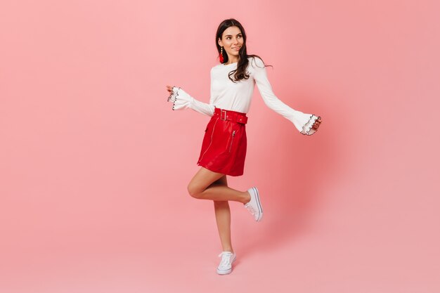 Full-length portrait of young dark-haired girl in leather red skirt and light blouse dancing on pink background.