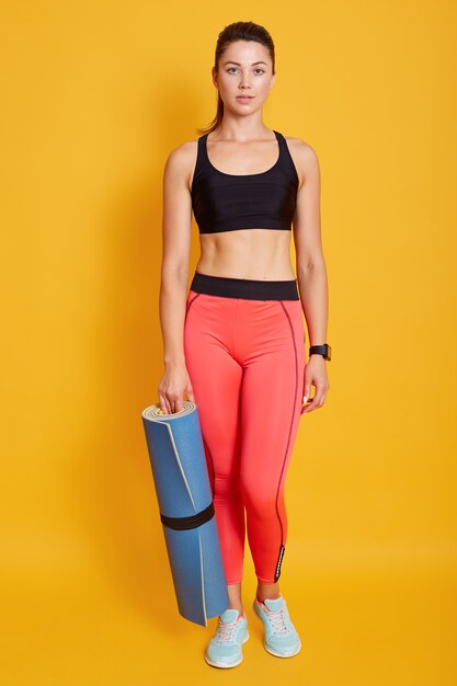 Full length portrait of young female with blue exercising mat in hand, posing isolated over yellow background