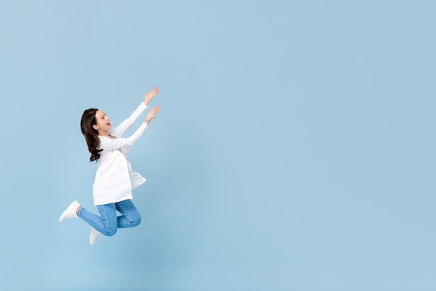 Full length portrait of young happy asian woman floating in mid-air with hands opening to copyspace beside in blue isolated wall