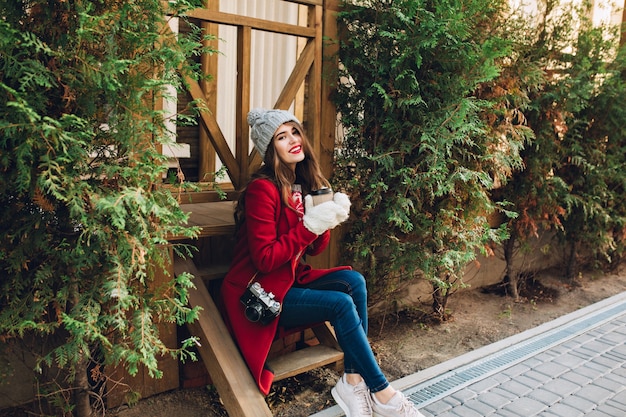 無料写真 赤いコート、ニット帽子、屋外の緑の枝の間の木製の階段の上に座っている白い手袋で完全な長さのかわいい女の子。彼女はコーヒーを持ち歩き、笑っています。