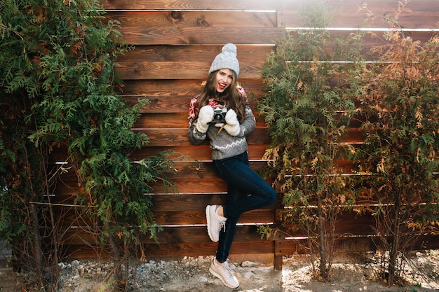 Full-length pretty girl with long hair in knitted hat and gloves holding camera on wooden .