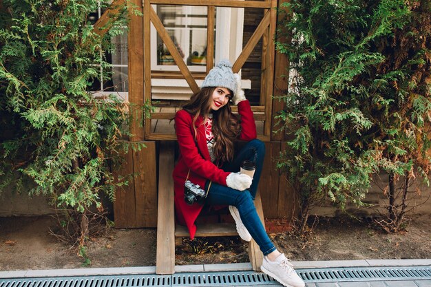 Full length pretty girl with long hair in red coat and knitted hat sitting on wooden stairs outdoor. She holds camera and coffee in white gloves, smiling .