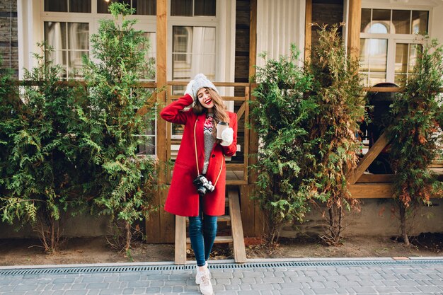 Full length pretty girl with long hair in red coat and knitted hat standing on wooden house . She holds camera and coffee to go in white gloves. Looks satisfied with closed eyes.