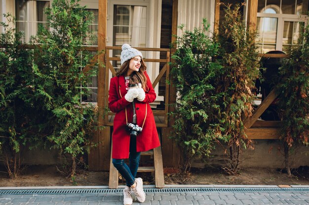 빨간 코트와 니트 모자 서 목조 주택에 긴 머리를 가진 전체 길이 예쁜 여자. 그녀는 카메라와 커피를 들고 흰 장갑을 끼고 옆으로 웃고 있습니다.