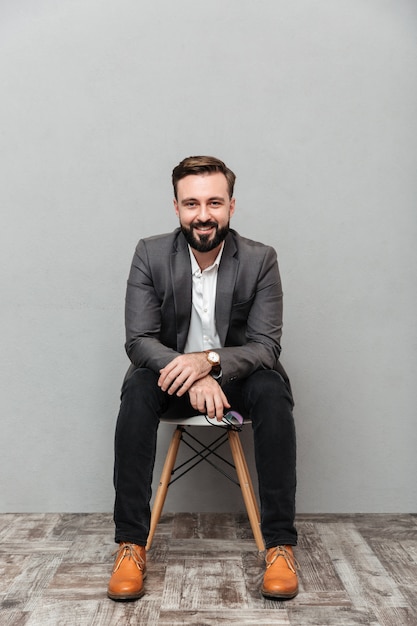 Free photo full-length relaxed bearded man sitting on chair in office, and smiling on camera isolated over gray