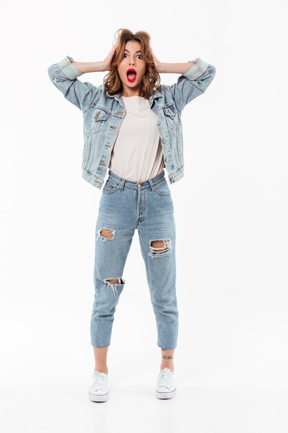 Full length Shocked woman in denim clothes holding her head  over white wall