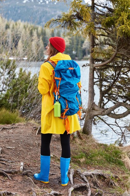 Full length shot of active female hiker stands on hill near mountain lake, wears red hat