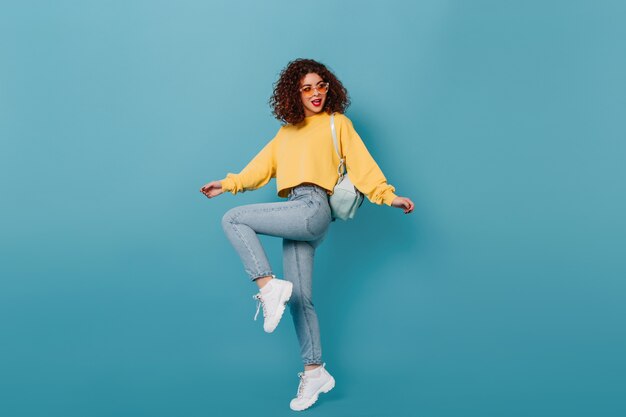 Full-length shot of active girl in skinny jeans and yellow sweatshirt raising her leg on blue space.