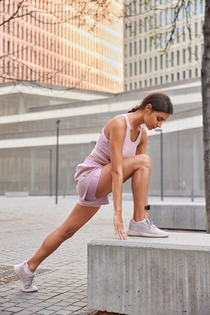 활동적인 근육질 여성의 전체 길이 샷은 운동복을 입은 콘크리트 돌에 기대어 운동을 하기 전에 도시 배경에 대해 야외에서 포즈를 취합니다. 운동 여성 모델은 달리기를 준비합니다