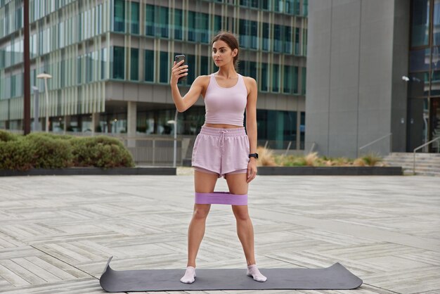 Full length shot of active slim woman records video for her sport blog dressed in tracksuit trains with resistance band poses on fitness mat in urban setting makes video call while having aerobics