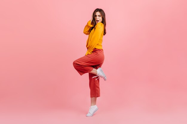 Full-length shot of amazed brunette on pink wall. Woman in bright blouse and pants coquettishly raised her leg.