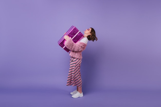 Full-length shot of amazing birthday girl in white sneakers holding her present.  curly female model in pink jacket posing with gift.