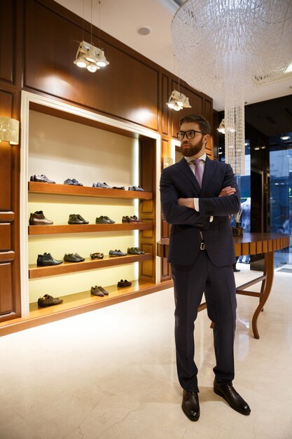 Full-length shot of bearded man in suit and glasses standing in wardrobe and looking away