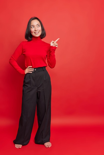 Full length shot of beautiful Asian woman in turtleneck and black trousers stands bare feet indicates away on blank copy space shows advertisement or promo attracts your attention to something