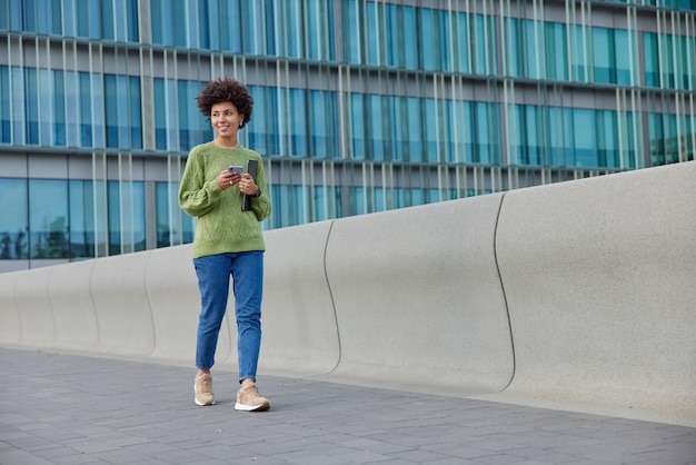 아름다운 곱슬머리 여성의 전체 길이 사진은 녹색 점퍼 청바지와 운동화를 신고 도시 비즈니스 센터 근처 외부에서 현대적인 장치를 가지고 산책하며 즐거운 표정을 짓습니다.