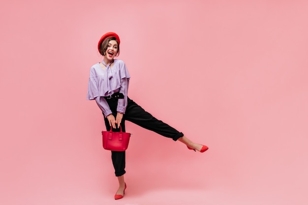 Foto gratuita colpo integrale di bella ragazza in pantaloni neri, top viola e berretto. donna sorridente, tenendo la borsa e alzando la gamba su sfondo rosa.