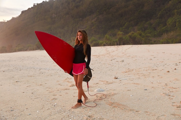 Full length shot of beautiful girl surfer dressed in boardshorts and black waterproof top