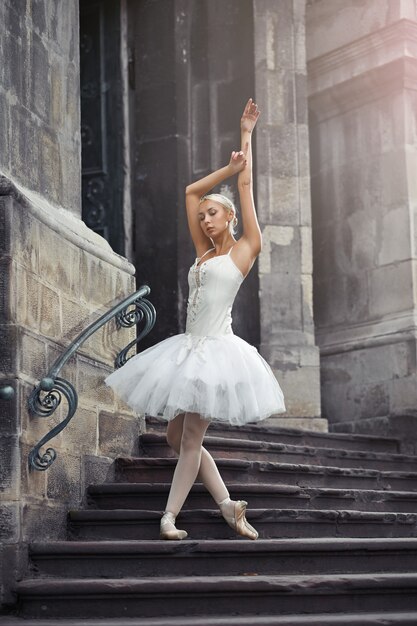 Снимок в полный рост красивой молодой артистки балета, изящно позирующей на лестнице старого здания в городе.