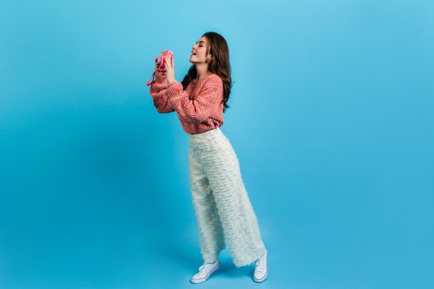 Full-length shot of charming brunette in white pants and pink sweater. Girl with interest takes picture on Instax.