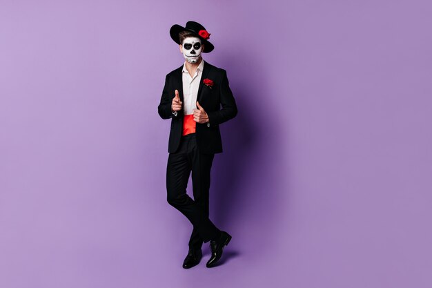 Full-length shot of charming guy with red satin belt under black jacket, posing in Halloween costume.