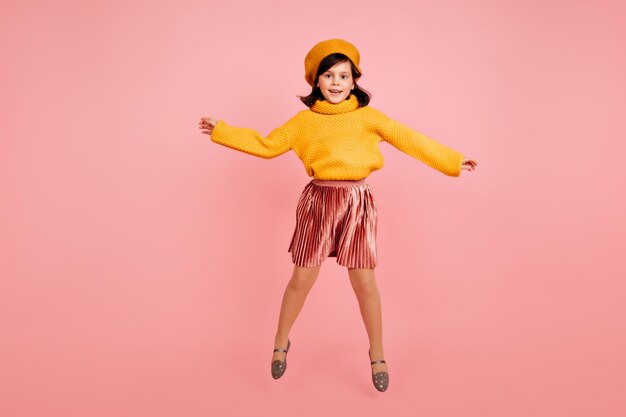 ピンクの背景にジャンプする興奮したプレティーンの女の子の黄色いベレー帽スタジオ写真のかわいい女性の子供のフルレングスのショット