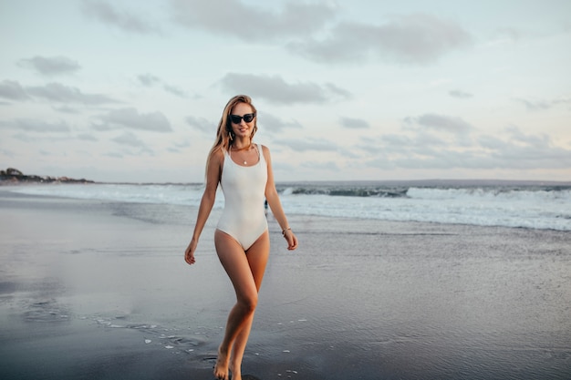 Full-length shot of enthusiastic woman in trendy swimsuit standing at ocean coast. Charming tanned lady in white swimwear posing with pleasure at seascape.