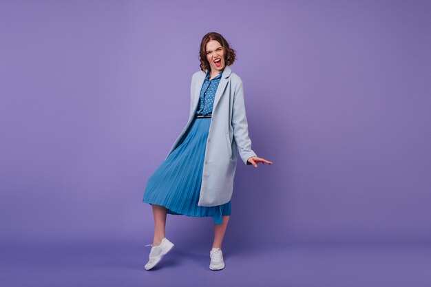 Full-length shot of excited curly woman in long dress making funny faces on purple wall. Winsome brunette girl posing in blue coat and smiling.