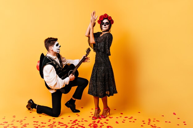 Full-length shot of extraordinary creative couple dancing and singing on orange wall. Girl and boy with skull masks posing