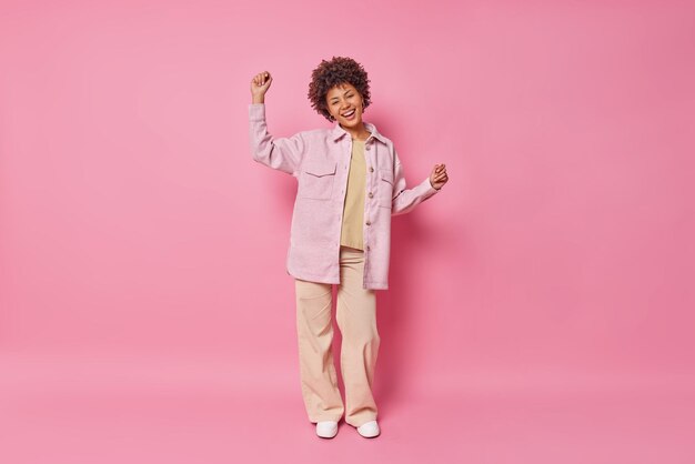 Full length shot of fashionable curly haired woman has fun dances carefree keeps arms raised wers stylish shirt trousers and sneakers poses against pink wall