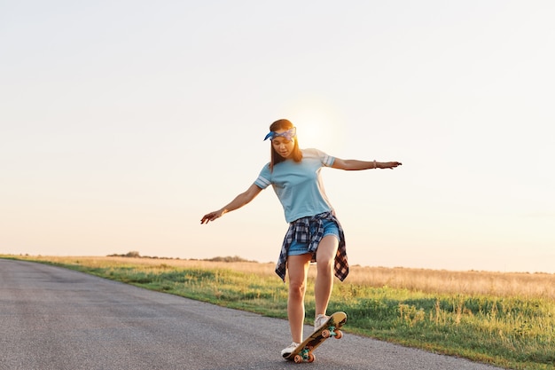 Full length shot of girl wearing casual attire skateboarding on empty street, spreading hands aside, enjoying riding, having concentrated facial expression.