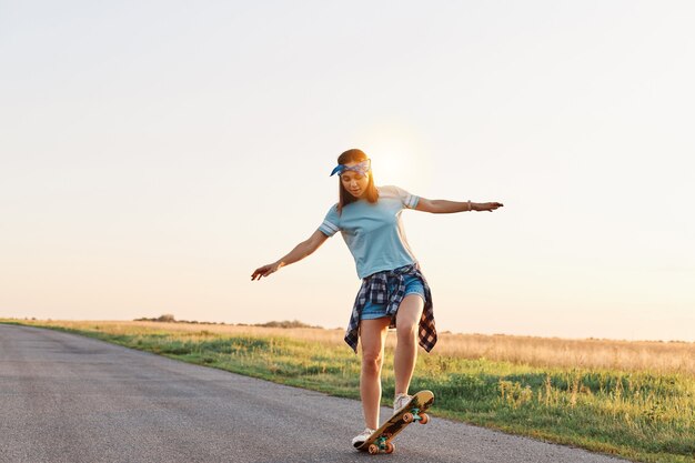 Снимок в полный рост девушки в повседневной одежде, катающейся на скейтборде по пустой улице, разводящей руки в сторону, наслаждающейся катанием, с сосредоточенным выражением лица.