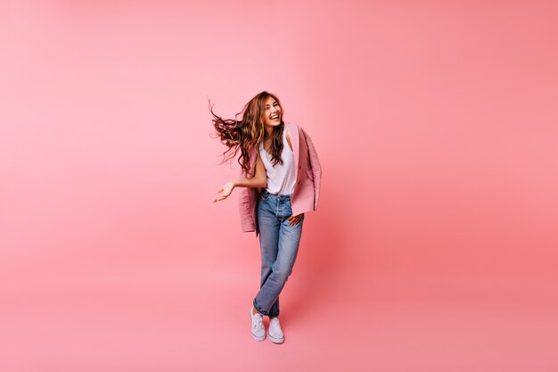 Full-length shot of gorgeous dark-haired lady standing in confident pose. Indoor portrait of pleased red-haired girl in pink jacket and jeans.