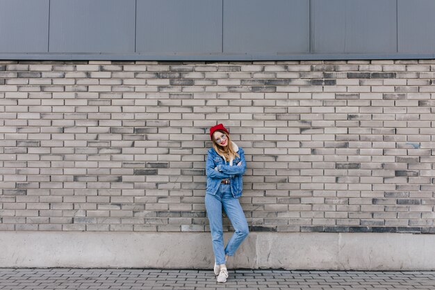 Full-length shot of gorgeous woman in white sneakers posing on urban wall with arms crossed. Outdoor portrait of attractive female model in red hat standing in front of brick wall.