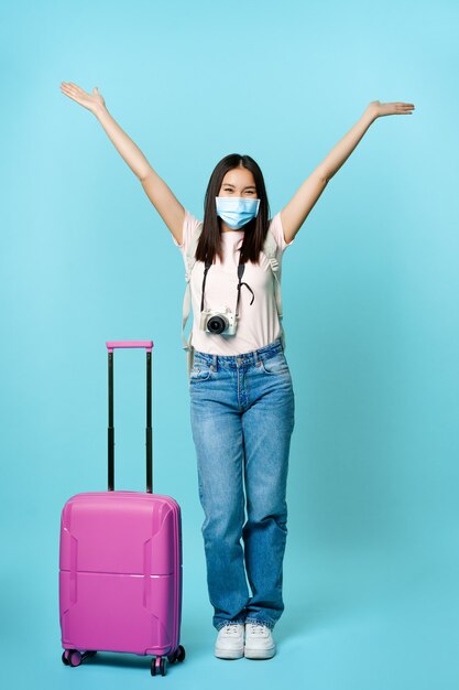 Снимок в полный рост счастливой азиатской девушки-туриста в медицинской маске, стоящей возле милого чемодана, идущей в отпуск во время пандемии covid, взволнованной поездкой за границу.